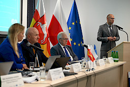 Przedstawiciel Komisji Europejskiej wypowiada się do mikrofonu za ambony. Wyświetla prezentację na ekranie rzutnika. Obok widać siedzących za stołem przewodniczących posiedzenia.