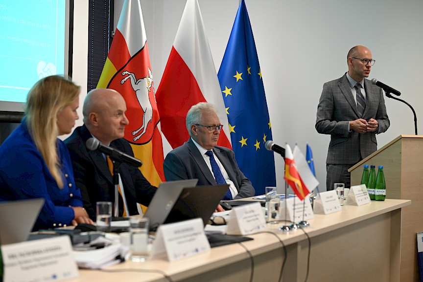 Przedstawiciel Komisji Europejskiej wypowiada się do mikrofonu za ambony. Wyświetla prezentację na ekranie rzutnika. Obok widać siedzących za stołem przewodniczących posiedzenia.