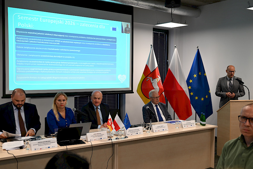 Przedstawiciel Komisji Europejskiej wypowiada się do mikrofonu zza ambony. Na ścianie na rzutniku wyświetlana jest prezentacja. 