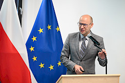 Przedstawiciel Komisji Europejskiej wypowiada się do mikrofonu. Ujęcie z bliska. Za nim widoczne są dwie flagi, Polski i Unii Europejskiej.