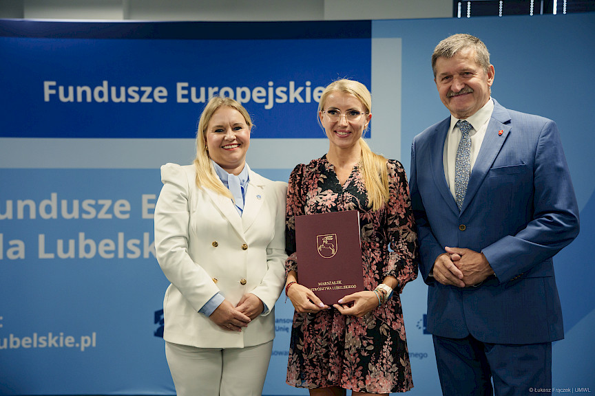 Dyrektor Departamentu Zarządzania Programami Regionalnymi Anna Brzyska stoi obok beneficjentki projektu, która trzyma bordową teczkę w rękach. Obok nich stoi mężczyzna.
