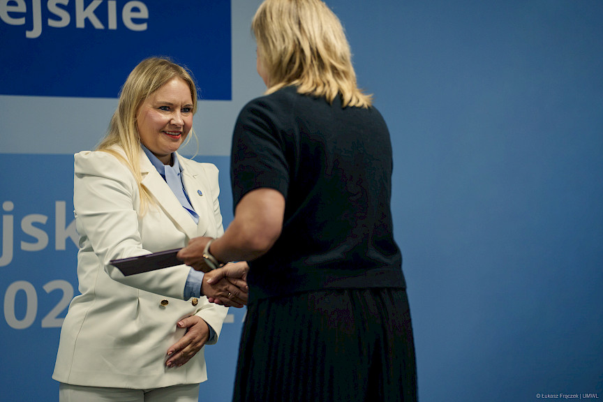 Dyrektor Departamentu Zarządzania Programami Regionalnymi Anna Brzyska podaje rękę w ramach gratulacji beneficjentce projektu, która odwrócona jest plecami do obiektywu.