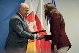 Marszałek Województwa Lubelskiego Jarosław Stawiarski podaje rękę w ramach gratulacji beneficjentce projektu. Za nimi znajdują się flagi.