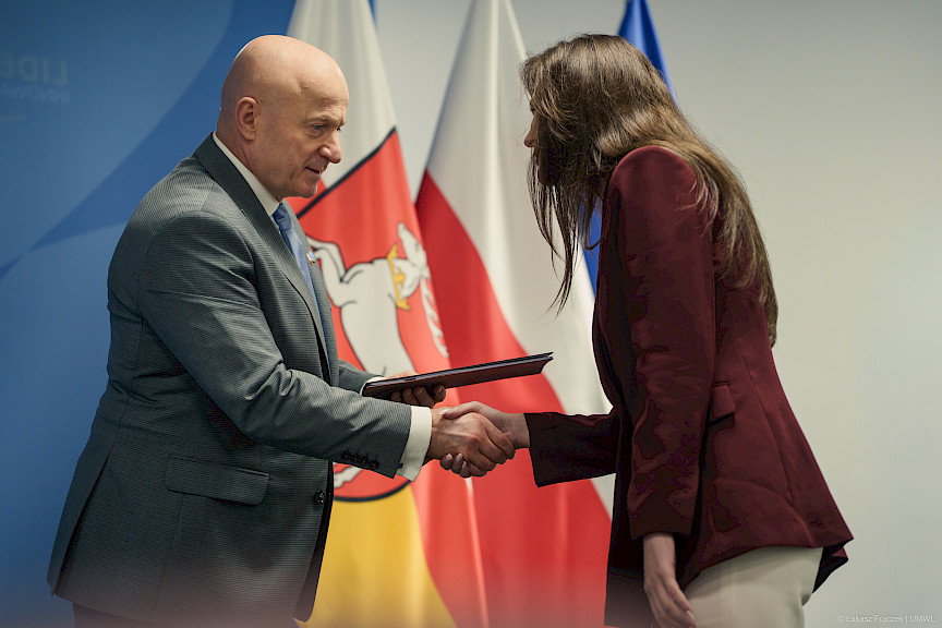 Marszałek Województwa Lubelskiego Jarosław Stawiarski podaje rękę w ramach gratulacji beneficjentce projektu. Za nimi znajdują się flagi.