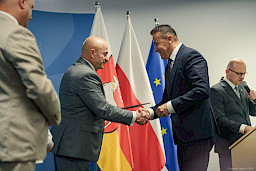 Marszałek Województwa Lubelskiego Jarosław Stawiarski podaje rękę w ramach gratulacji beneficjentowi projektu. W oddali widać prowadzącego wydarzenie, który stoi za amboną z mikrofonem.