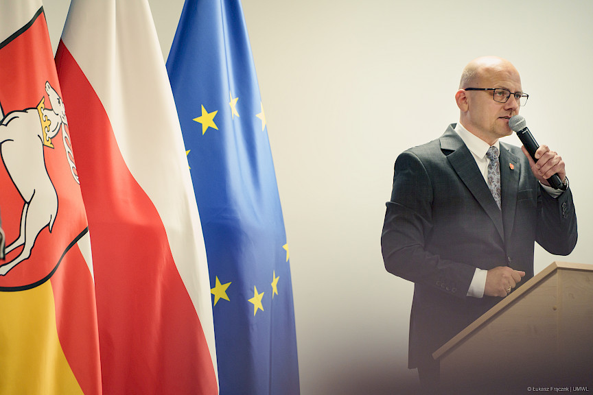 Zdjęcie prowadzącego wydarzenie, stoi przy ambonie z mikrofonem, obok widoczne są flagi, Polski, Województwa Lubelskiego oraz Unii Europejskiej.
