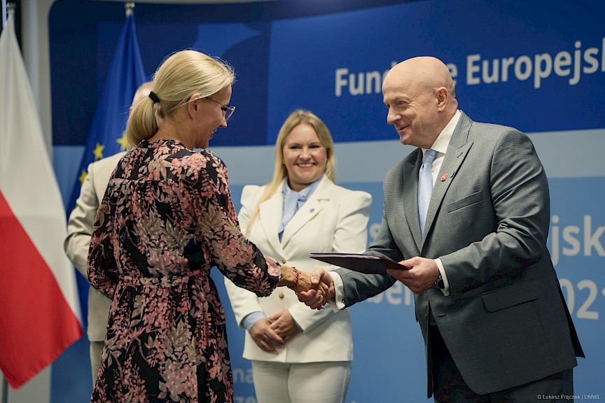 Marszałek Województwa Lubelskiego Jarosław Stawiarski podaje rękę w ramach gratulacji beneficjentce projektu. Za nimi widać Dyrektor Annę Brzyską, która szeroko uśmiecha się.