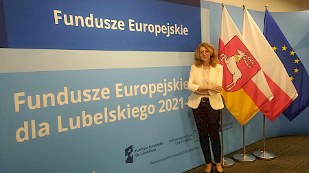 Prelegentka stoi przy ściance programu Fundusze Europejskie dla Lubelskiego 2021–2027, obok flag Województwa Lubelskiego, Polski i Unii Europejskiej.