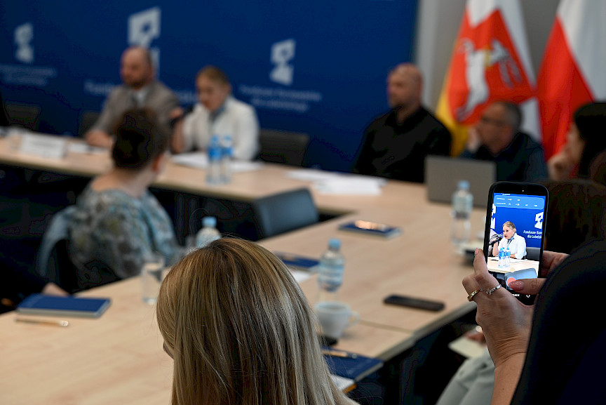 Ujęcie telefonu trzymanego przez inną osobę, która nagrywa Dyrektor Annę Brzyską. Na sali w sposób niewyraźny widoczni są uczestnicy szkolenia siedzący przy stołach.