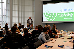 Zastępca Dyrektora Departamentu Zarządzania Programami Regionalnymi Piotr Budyńczuk przemawia przy ambonie do mikrofonu. Na sali siedzą uczestnicy spotkania. Na ekranie wyświetlany jest obraz z projektora dotyczący tematyki szkolenia.