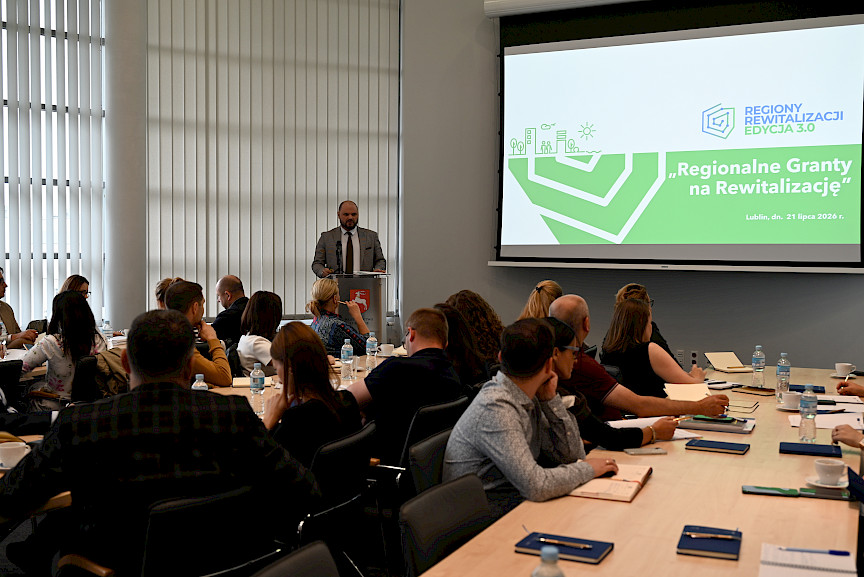 Zastępca Dyrektora Departamentu Zarządzania Programami Regionalnymi Piotr Budyńczuk przemawia przy ambonie do mikrofonu. Na sali siedzą uczestnicy spotkania. Na ekranie wyświetlany jest obraz z projektora dotyczący tematyki szkolenia.
