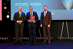 Marszałek Województwa Lubelskiego Jarosław Stawiarski oraz Wicemarszałek Marek Wojciechowski stoją z beneficjentem projektu, który trzyma bordową teczkę w rękach.