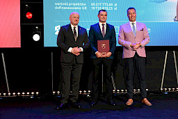 Marszałek Województwa Lubelskiego Jarosław Stawiarski z Członkiem Zarządu Marcinem Szewczakiem stoją z beneficjentem projektu, który trzyma bordową teczkę w rękach..