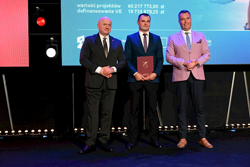 Marszałek Województwa Lubelskiego Jarosław Stawiarski z Członkiem Zarządu Marcinem Szewczakiem stoją z beneficjentem projektu, który trzyma bordową teczkę w rękach..