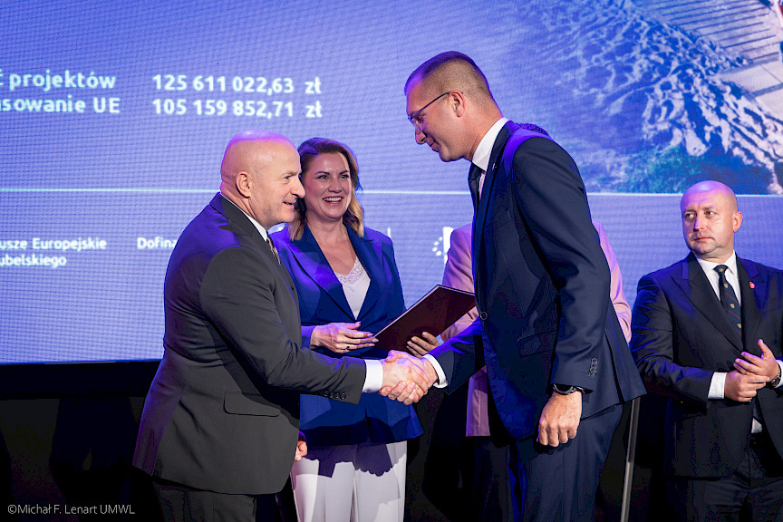 Marszałek Województwa Lubelskiego Jarosław Stawiarski podaje rękę beneficjentowi projektu w ramach gratulacji. Za nimi stoi uśmiechnięta kobieta oraz Wicemarszałek Piotr Breś.