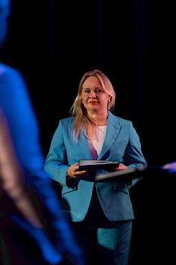 Dyrektor Departamentu Zarządzania Programami Regionalnymi Anna Brzyska w niebieskim garniturze stoi z teczką, uśmiecha się i patrzy na inną osobę.