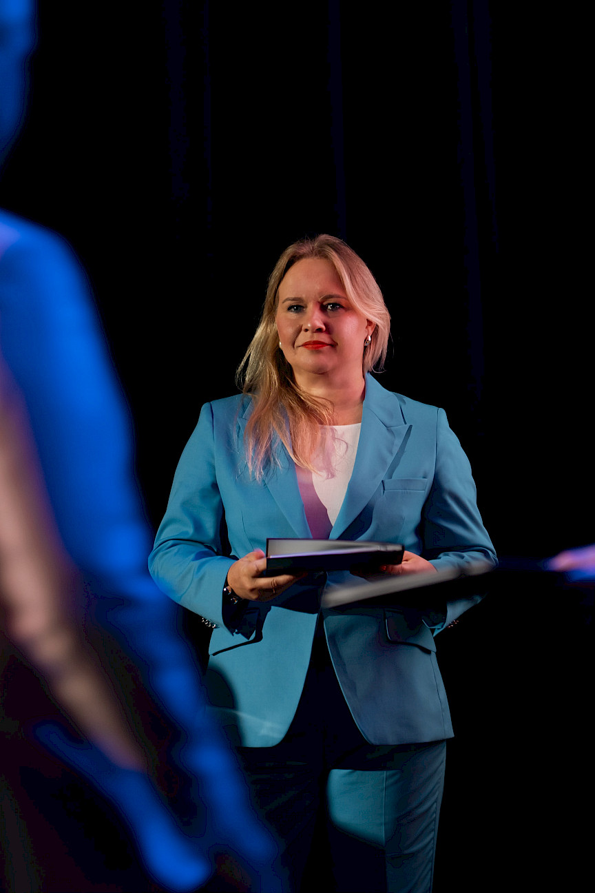 Dyrektor Departamentu Zarządzania Programami Regionalnymi Anna Brzyska w niebieskim garniturze stoi z teczką, uśmiecha się i patrzy na inną osobę.