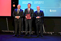 Marszałek Województwa Lubelskiego Jarosław Stawiarski i Wicemarszałek Piotr Breś stoją z beneficjentem projektu, który trzyma bordową teczkę z umową.
