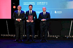Marszałek Województwa Lubelskiego Jarosław Stawiarski i Wicemarszałek Piotr Breś stoją z beneficjentem projektu, który trzyma bordową teczkę z umową.