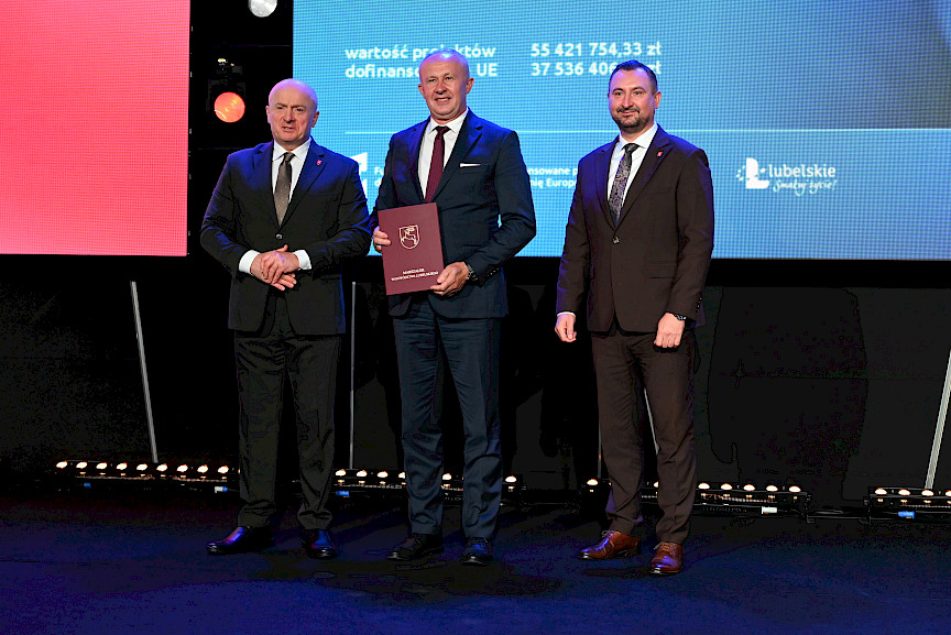 Marszałek Województwa Lubelskiego Jarosław Stawiarski z Wicemarszałkiem Markiem Wojciechowskim stoją z beneficjentem, który trzyma bordową teczkę w umową.
