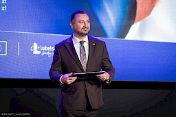 Wicemarszałek Marek Wojciechowski trzyma w rękach teczkę z umową i czeka na podejście beneficjenta.