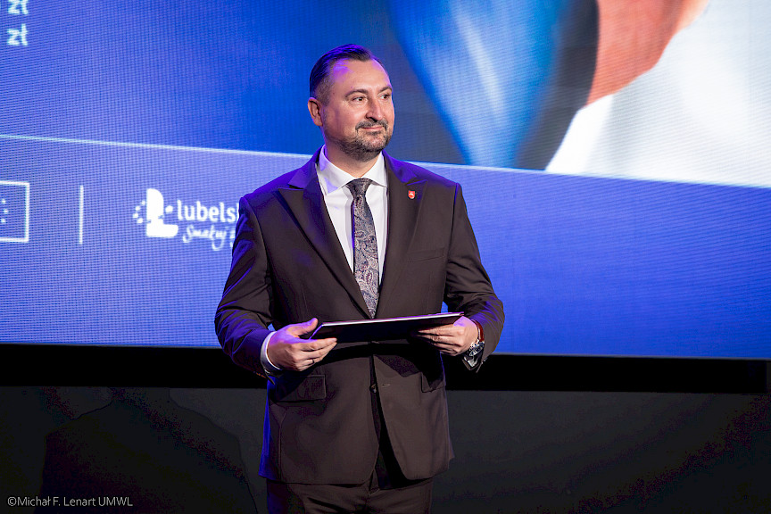 Wicemarszałek Marek Wojciechowski trzyma w rękach teczkę z umową i czeka na podejście beneficjenta.