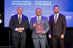 Marszałek Województwa Lubelskiego Jarosław Stawiarski z Wicemarszałkiem Markiem Wojciechowskim stoją z beneficjentem projektu, który trzyma bordową teczkę z umową.
