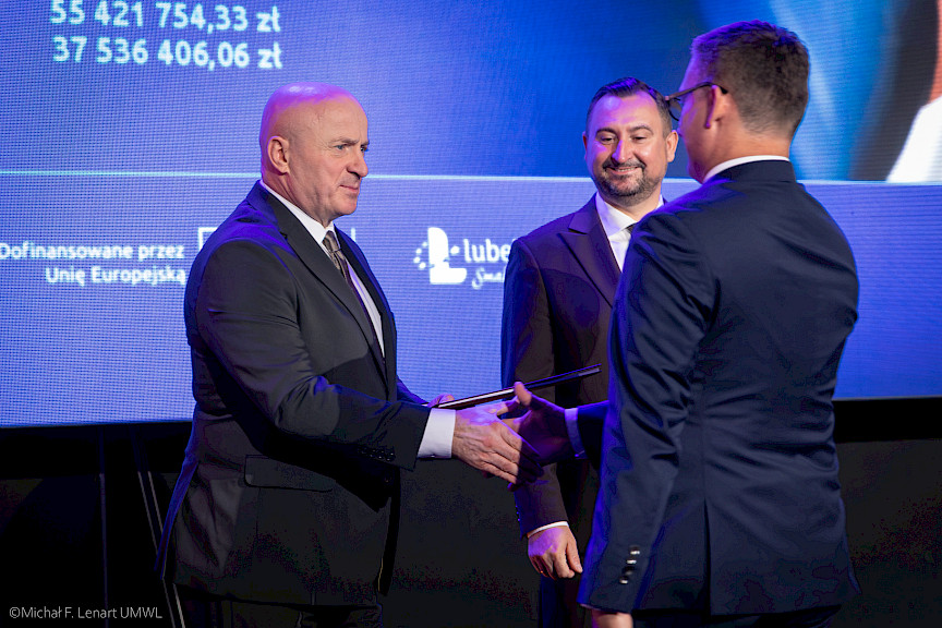 Marszałek Województwa Lubelskiego Jarosław Stawiarski podaje rękę beneficjentce projektu w ramach gratulacji. Obok nich stoi i uśmiecha się Wicemarszałek Marek Wojciechowski.