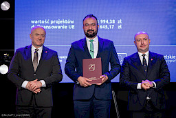 Marszałek Województwa Lubelskiego Jarosław Stawiarski z Wicemarszałkiem Piotrem Bresiem stoją z beneficjentem projektu, który trzyma bordową teczkę z umową.