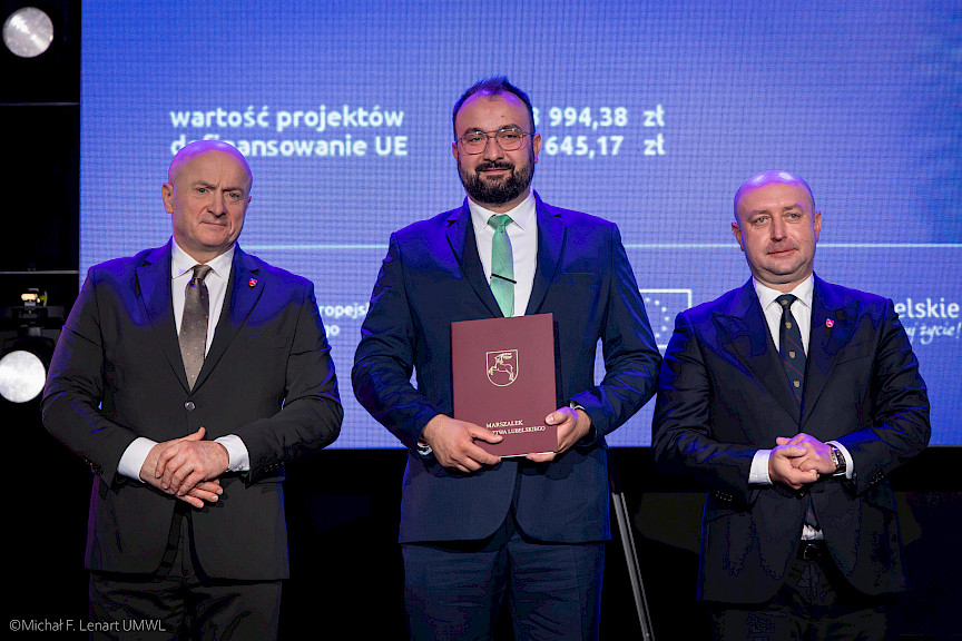 Marszałek Województwa Lubelskiego Jarosław Stawiarski z Wicemarszałkiem Piotrem Bresiem stoją z beneficjentem projektu, który trzyma bordową teczkę z umową.