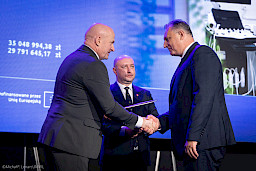 Marszałek Województwa Lubelskiego Jarosław Stawiarski podaje rękę beneficjentowi projektu w ramach gratulacji oraz podaje mu teczkę w umowa. Za nimi stoi Wicemarszałek Piotr Breś.