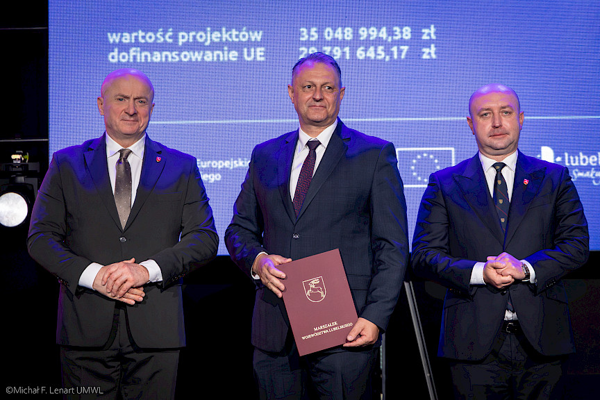 Marszałek Województwa Lubelskiego Jarosław Stawiarski oraz Wicemarszałek Piotr Breś stoją z beneficjentem projektu, który trzyma bordową teczkę w umową.