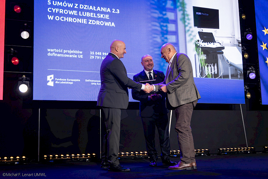 Marszałek Województwa Lubelskiego Jarosław Stawiarski podaje rękę beneficjentowi projektu w ramach gratulacji. Obok nich stoi Wicemarszałek Piotr Breś.