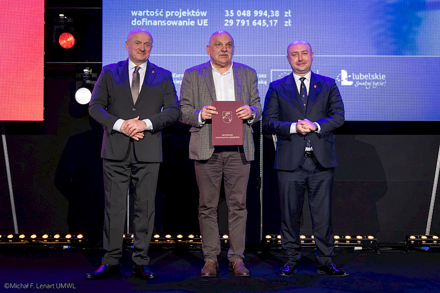 Marszałek Województwa Lubelskiego Jarosław Stawiarski oraz Wicemarszałek Piotr Breś stoją z beneficjentem projektu, który trzyma bordową teczkę w umową.