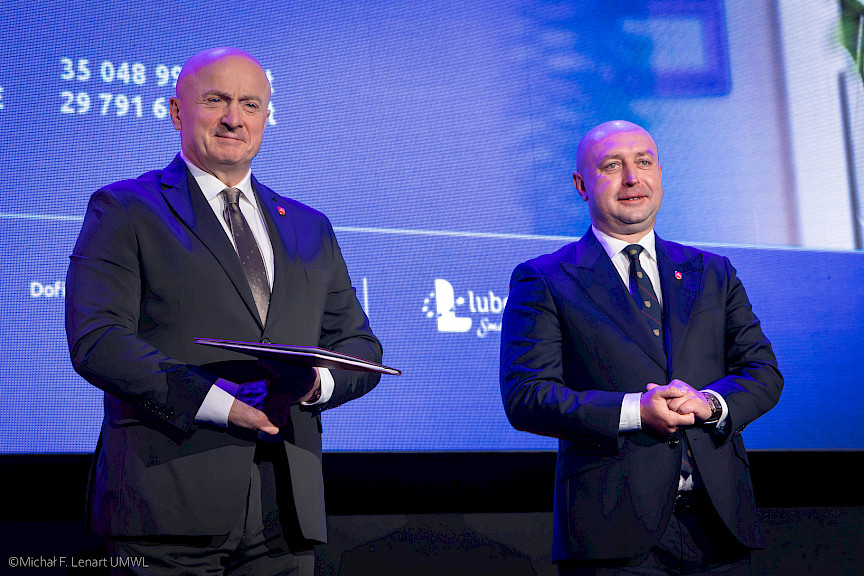 Marszałek Województwa Lubelskiego Jarosław Stawiarski oraz Wicemarszałek Piotr Breś stoją i czekają na podejście beneficjenta projektu aby wręczyć mu umowę.