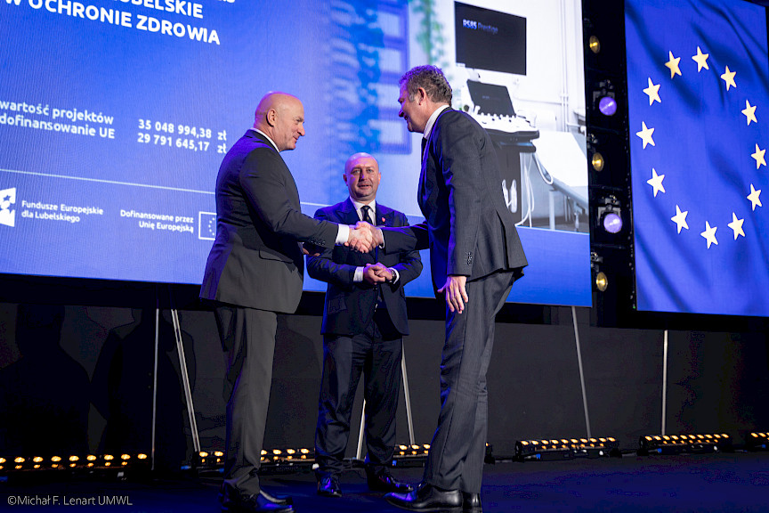 Marszałek Województwa Lubelskiego Jarosław Stawiarski podaje rękę beneficjentowi projektu w ramach gratulacji. Obok nich stoi Wicemarszałek Piotr Breś.