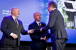Marszałek Województwa Lubelskiego Jarosław Stawiarski podaje teczkę z umową o dofinansowanie beneficjentowi projektu. Obok nich stoi Wicemarszałek Piotr Breś i podaje rękę beneficjentowi.