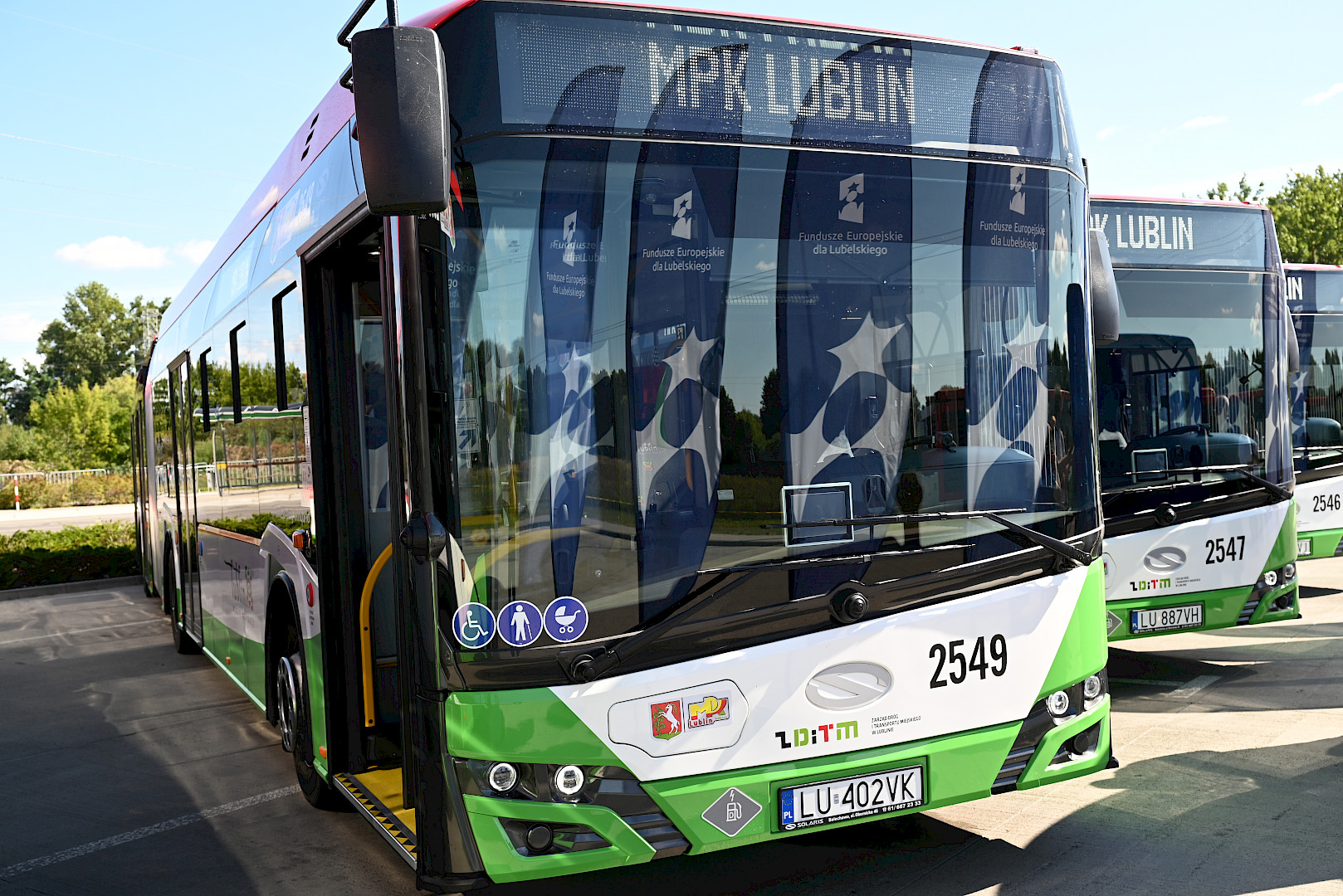 Ujęcie przodu autobusu. Na górze szyby wyświetla się napis MPK Lublin. Autobus jest w kolorach zielono, biało, czarnych. Obok niego w widoczne są jeszcze dwa autobusy ustawione obok siebie.