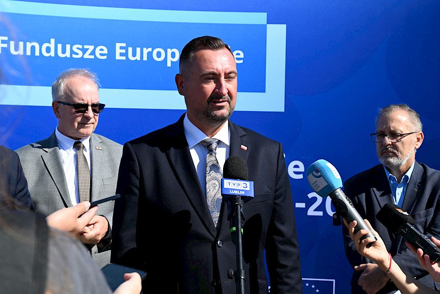 Wicemarszałek Województwa Lubelskiego Marek Wojciechowski udziela wywiadu mediom do mikrofonów. Za nim stoi dwóch mężczyzn.