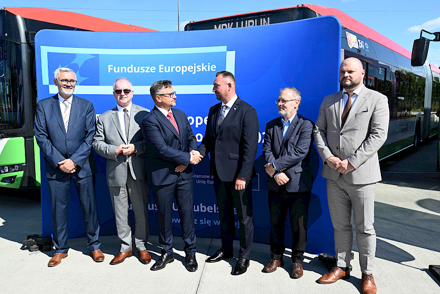 Wicemarszałek Województwa Lubelskiego Marek Wojciechowski podaje rękę Prezesowi MPK Lublin Bogdanowi Kołciukowi na tle niebieskiej ścianki promującej Fundusze Europejskie. Obok nich po obu stronach stoją inni mężczyźni.