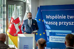Mężczyzna w garniturze  przemawia do mikrofonu do uczestników siedzących na sali. Za nim znajduje się niebieska flaga Unii Europejskiej, Polski i Województwa Lubelskiego oraz niebieska ścianka promująca fundusze europejskie.