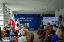 Kobieta w białym fartuchu przemawia do mikrofonu do uczestników siedzących na sali.. Na monitorze wyświetla obraz z informacjami dotyczącymi szpitala. Za nią stoi niebieska ścianka promująca fundusze europejskie.