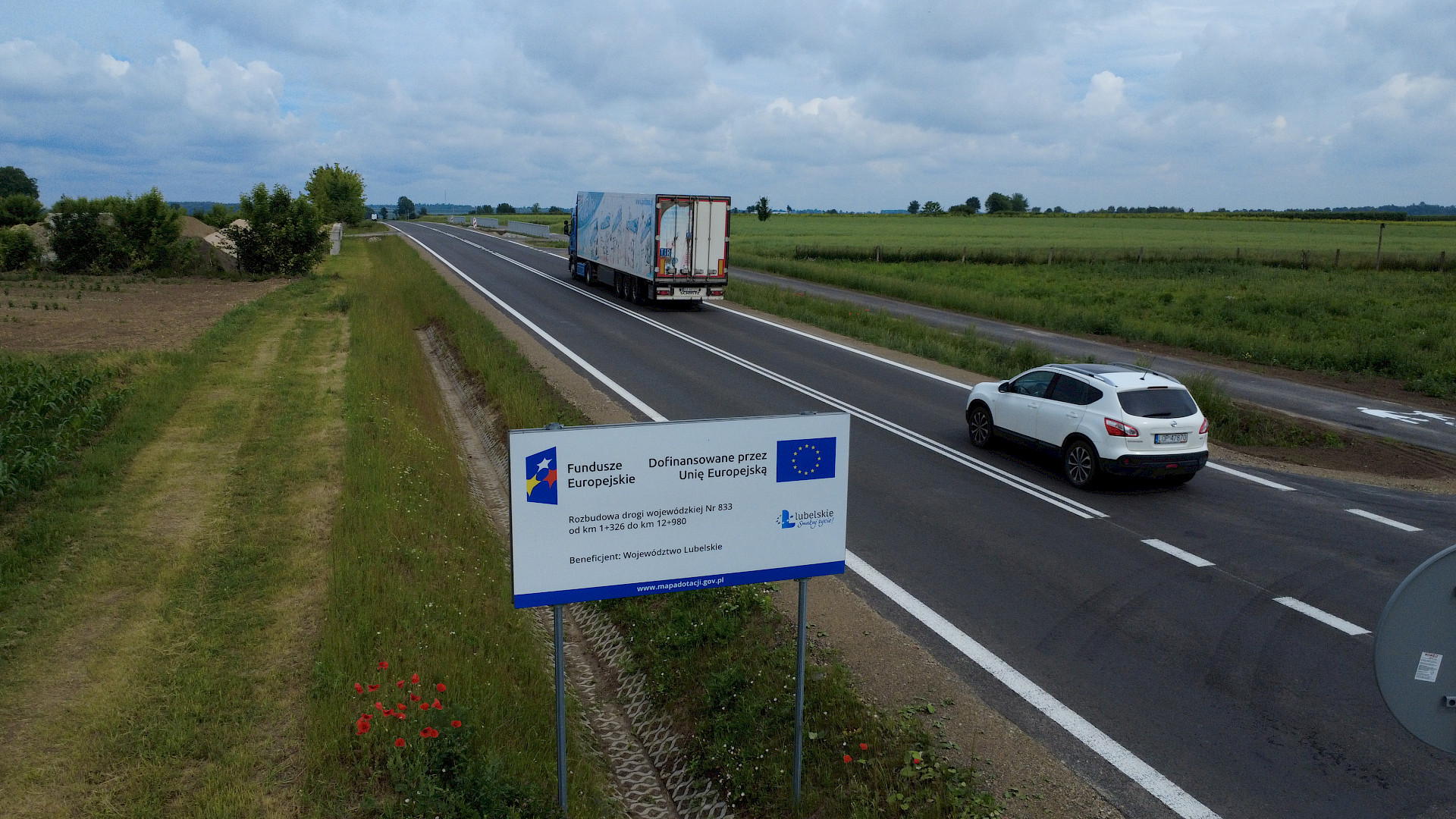 Ulica z dwoma jadącymi samochodami. W koło widoczne są zielone tereny. Na pierwszym planie, przy drodze stoi tablica informacyjna dotycząca dofinansowania z funduszy europejskich.