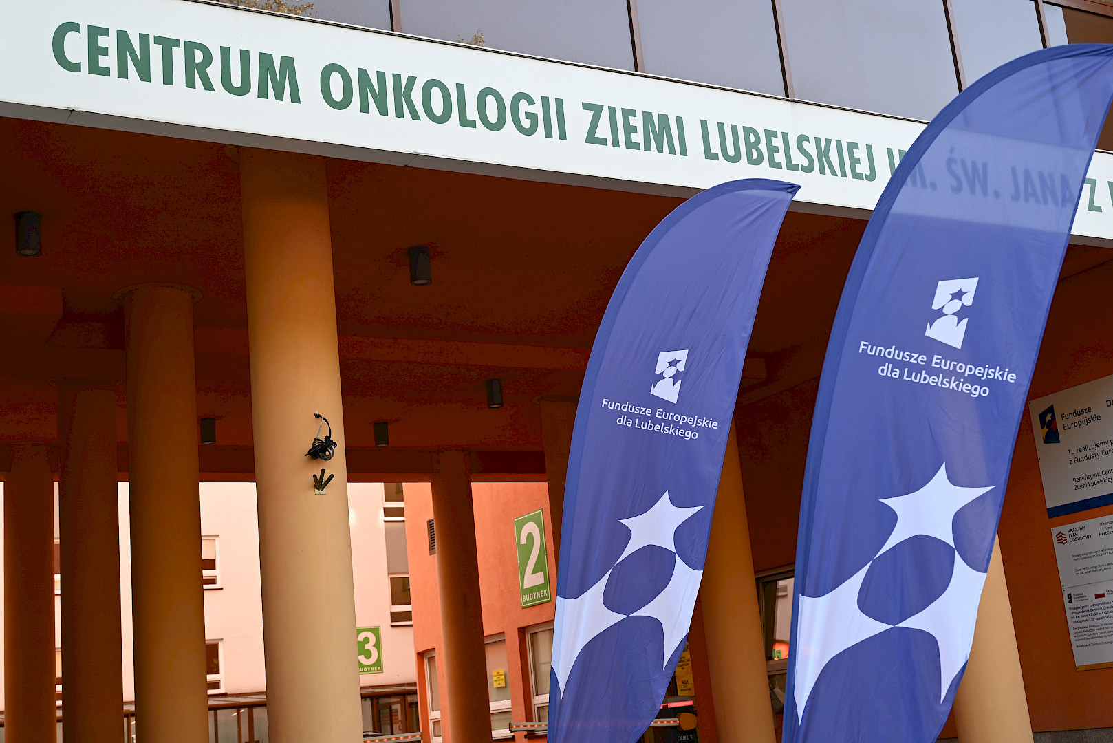 Fragment budynku szpitala. Na górze widnieje napis Centrum Onkologii Ziemi Lubelskiej, poniżej napisu stoją dwie granatowe flagi promujące fundusze europejskie dla lubelskiego.