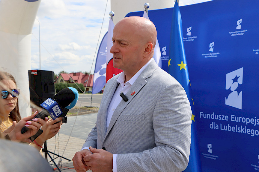Marszałek Województwa Lubelskiego Jarosław Stawiarski udziela wywiadu mediom do mikrofonów. Za nim widoczna jest ścianka promująca fundusze europejskie.