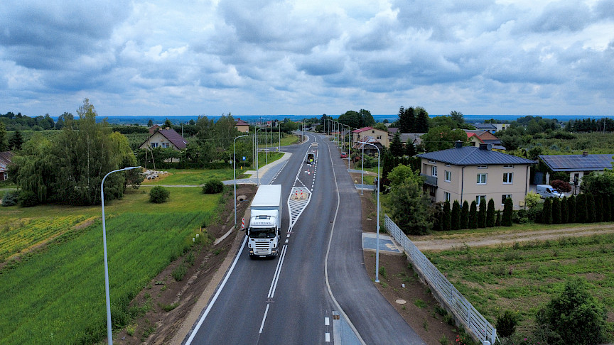 Ujęcie z lotu ptaka na drogę w koło widoczne są zielone tereny, budynki oraz jadące samochody na drodze.
