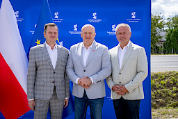Marszałek Województwa Lubelskiego Jarosław Stawiarski pozuje do zdjęcia z dwoma mężczyznami w garniturach. Za nimi stoi niebieska ścianka promująca  fundusze europejskie dla lubelskiego.