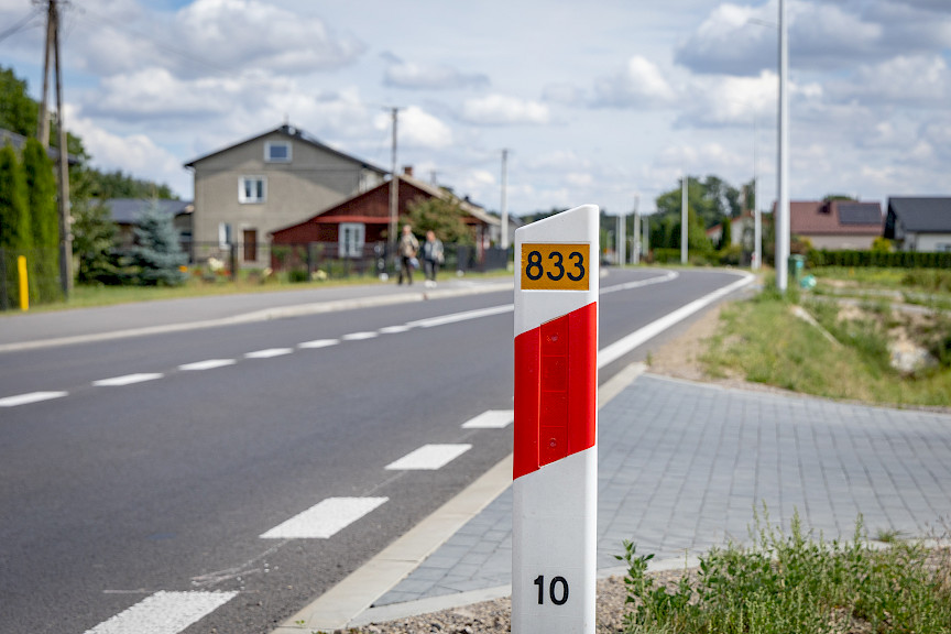 Zdjęcie fragmentu drogi, w koło widoczne są budynki. Na pierwszym planie widnieje słupek z numerem drogi 833.