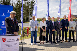 Burmistrz Urzędowa Paweł Dąbrowski wypowiada się do mikrofonu do uczestników wydarzenia. Obok stoją mężczyźni w garniturach oraz jedna kobieta. Za nimi stoją niebieskie flagi promujące fundusze europejskie dla lubelskiego