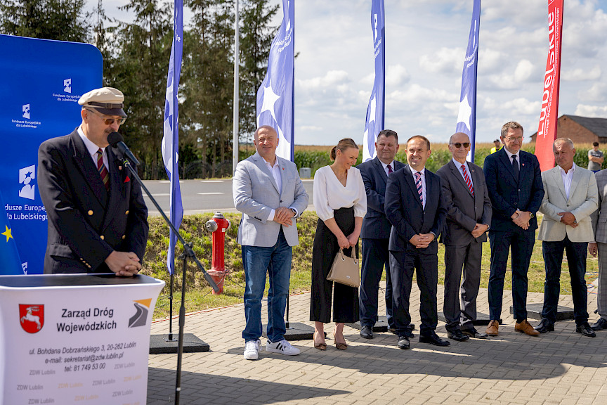 Burmistrz Urzędowa Paweł Dąbrowski wypowiada się do mikrofonu do uczestników wydarzenia. Obok stoją mężczyźni w garniturach oraz jedna kobieta. Za nimi stoją niebieskie flagi promujące fundusze europejskie dla lubelskiego